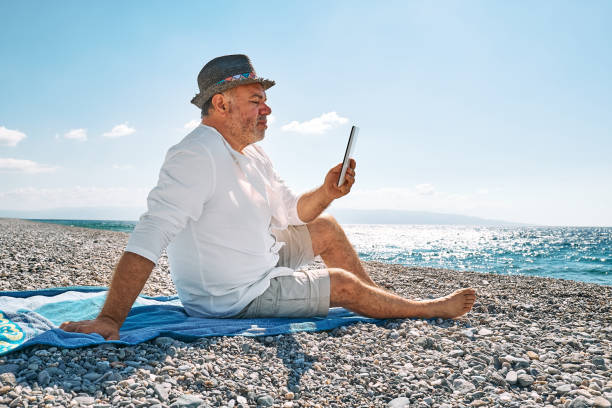 ビーチで休息し、電子タブレットで電子書籍を読んでいる中年のあごひげを生やした男性。夏休みに読書とリラックスをコンセプトに。ビーチ文学 - reading beach e reader men ストックフォトと画像