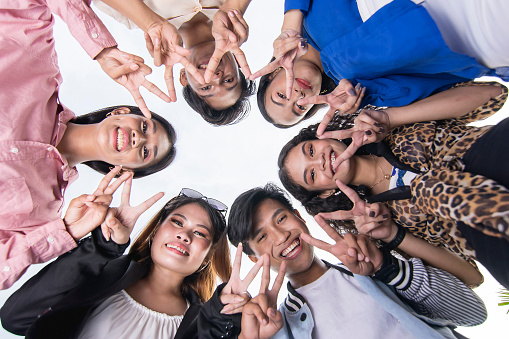 A team of six young asian friends huddle together, looking down and making double peace signs. Perspective of camera looking up.