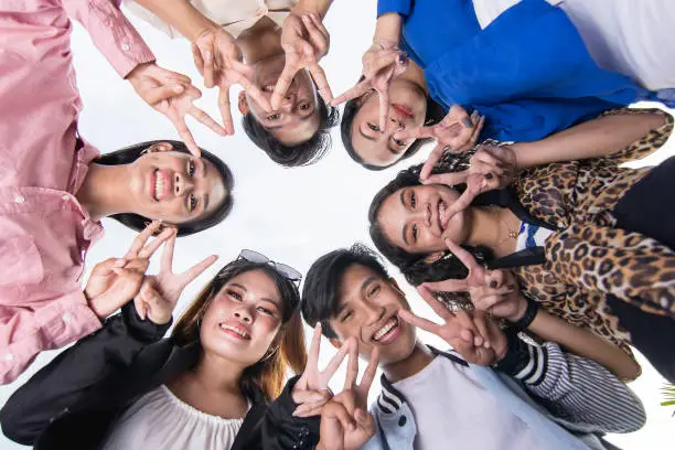 Photo of A team of six young asian friends huddle together, looking down and making double peace signs. Perspective of camera looking up.