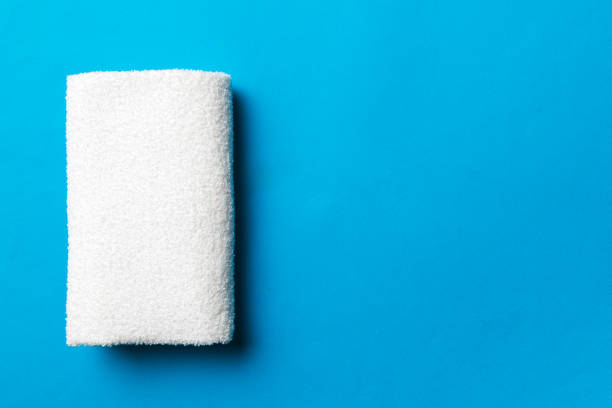 This is a close up photo of a cobalt blue, terry cloth towel, folded to depict a rose bloom.  The flower is centered in the photo, turned at a very slight angle to the right.  The background is stark white.  The edges of the towel appear to blur against the white background. 