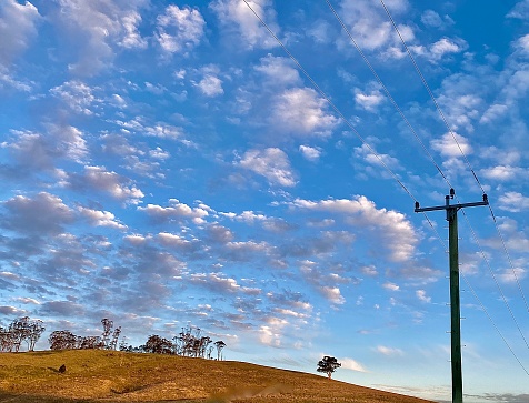 Horizontal country landscape of wood post power lines with background dry brown hill with cattle and sparse trees under a cloudy sky in rural Australia