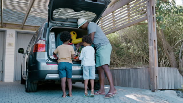 Family unpacking road trip suitcases, beach holiday inflatable toys from a car at resort. Adorable little sons helping single father prepare for vacation abroad. Man, children getting ready to travel