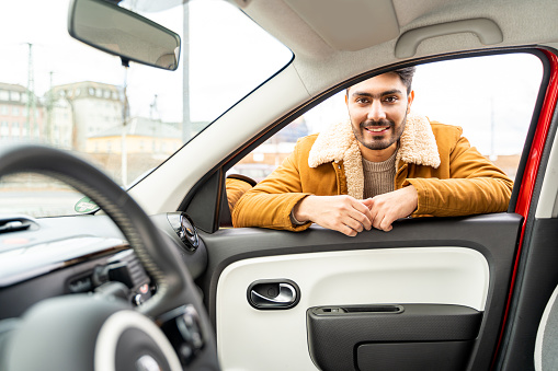Smiling spanish or arab eastern ethnicity cheerful man in yellow jacket leaned on door looking through window of car as passanger asking for ride or driver. Travel, exam, lesson, learning, taxi driver