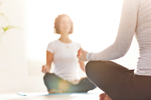 Two women meditating on a yoga mat with copy space. Young people sitting on the floor practicing mediation by doing the lotus pose feeling calm and at peace inside a bright and quiet wellness center