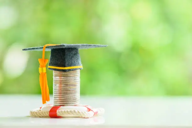 Photo of Tuition protection service and tuition refund insurance, financial concept : Black graduation cap or a mortarboard placed higher on top of a coin stack with a red lifebuoy on a table.