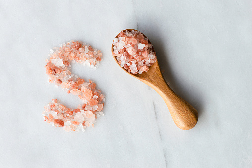 S written with pink Himalayan salt and a wooden spoon with a heap of Himalayan salt on white marble.