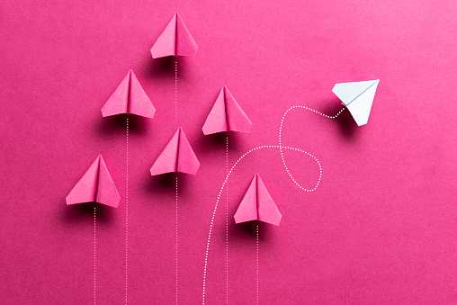 White paper plane is flying to the right on pink background where a group of pink paper planes are flying straight up.