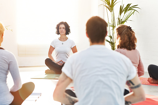 Woman meditating in lotus pose while teaching a class in a zen yoga studio. Group of yogis sitting together, finding inner peace and balance. Practising calming the mind with breathing exercises