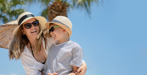 Portrait of young woman in beachwear embracing her son. Mother and kid enjoying summer vacation in tropical country with palm trees on background. Copy space.