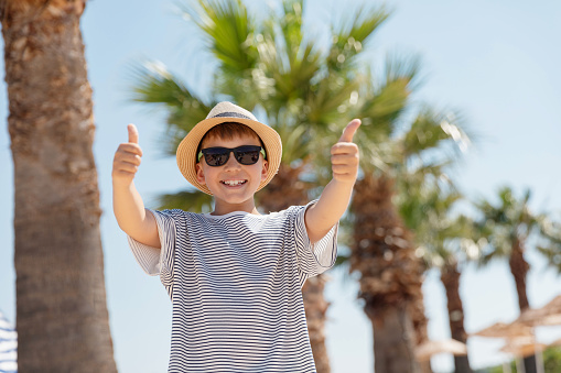 Portrait of smiling kid boy with in straw hat and sunglasses showing thumb up staying outdoor with palm trees and sky on the background looking at the camera.