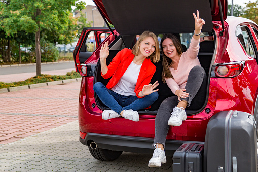 Two beautiful young smiling female friends showing free sign with fingers while sitting in open trunk of red modern car near travel baggage near green trees on a sunny summer day ready for a trip