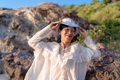 Portrait of a happy mature woman smiling outdoors. Happy smiling mature woman on the rock at sea beach, positive female wearing a white hat. Happiness, joy, relaxation, emotions, middle-aged people