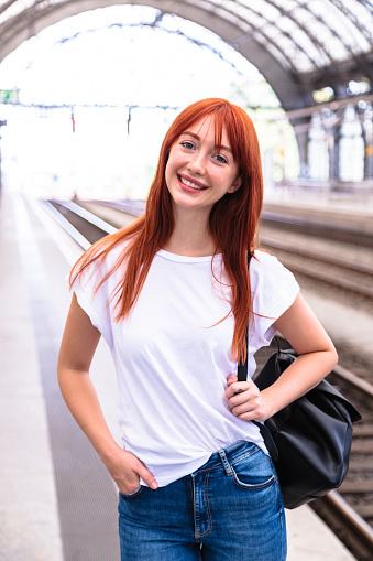 Portrait of young redhead smiling Caucasian woman traveler dressed casually with backpack standing near railroad alone on railway station waiting arrival of train at daytime. Travel and transport