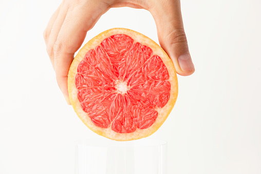 Human hand is about to put a grapefruit into a drinking glass on front of white background.