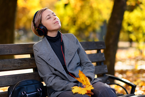 beautiful woman portrait, she is sitting on a bench in the autumn park and listens to music with headphones, trees with yellow leaves, bright sunny day