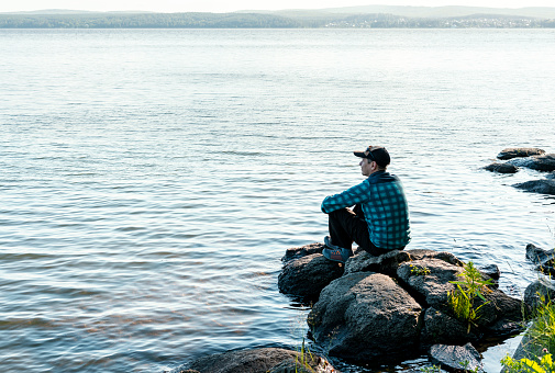 young man in a cap looking at the view of the lake or river sitting on the stones Enjoying nature landscape