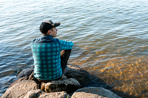 young man in a cap looking at the view of the lake or river sitting on the stones Enjoying nature landscape