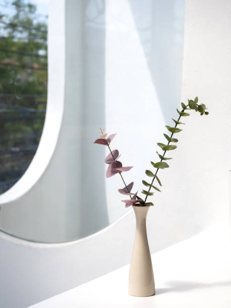 istock Home interior elegant floral vase decor, soft white composition. Beautiful pink and green leaves branches in white tall vase on white table and curved shape wall background, vertical, minimal style. 1407862095
