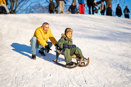 Young middle-aged cheerful smiling father in yellow jacket and jeans pushes wooden sled with his little african or latin son from crowdy snowy hill in park on sunny winter day. Holidays, parenthood