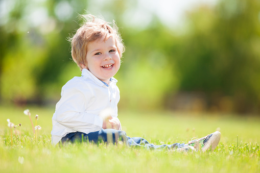 Cute smiling baby boy on the green meadow. Close up portrait of fun happy boy. Kid in the sitting on the green grass and looks at camera.
