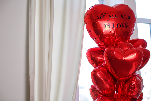 A garland of red balloons in the shape of a heart with the inscription All you need is love on the background of a window and a gray curtain