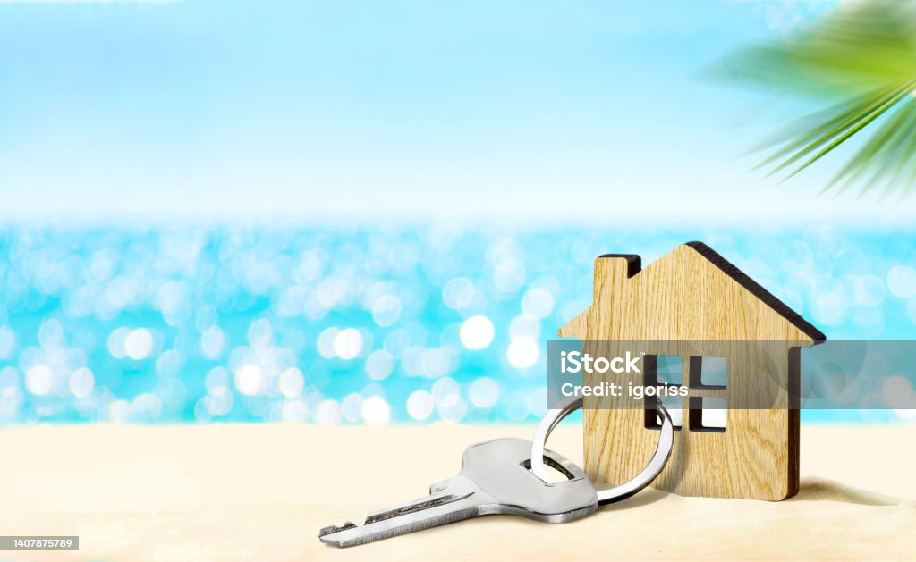 House by the sea concept. Figure of house and key on sandy beach Model of house and key on sandy beach with sea and sky on the background Vacation Rental Stock Photo