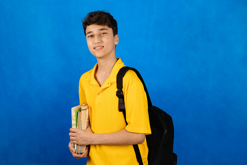 Cute teenager in a yellow T-shirt with a backpack and books on a blue background, back to school.