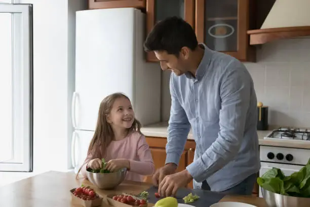 Little cute girl help to loving father with food preparation. Dad teach his 6s daughter cooking vegetable salad, cutting tomatoes cherry and talking, enjoy communication and cookery in kitchen at home