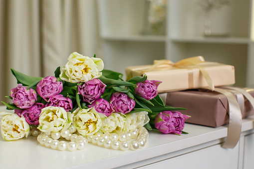 A bouquet of peony tulips of lemon and pink color lies on the table against the background of white pearls and two boxes with gifts