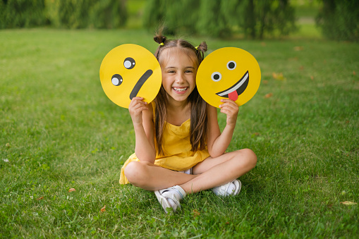 A laughing, cheerful child holds two emoticons in his hands - a sad, upset one and a smile face showing his tongue