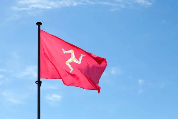Flag of Mann waving atop of its pole against a blue sky.