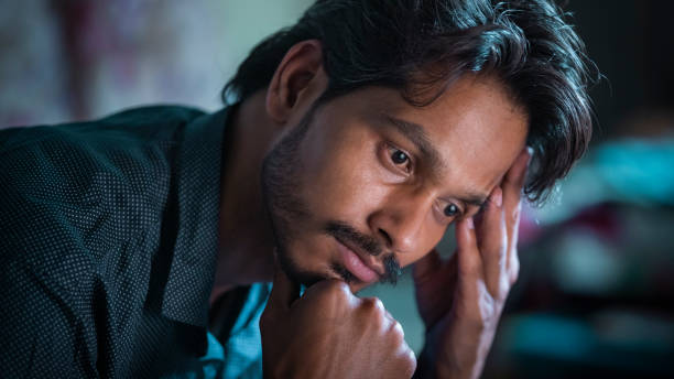 Sad and worried young man contemplates holding his head at home. An Asian/Indian sad single young man contemplates deeply holding his head at home in this indoor waist-up image with selective focus. head in hands photos stock pictures, royalty-free photos & images
