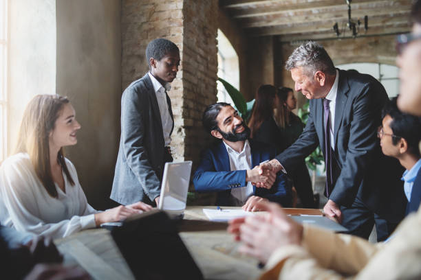 Two businessmen make a deal by shaking hands sitting at a table together with other multiracial employees - business lifestyle and agreements concept stock photo