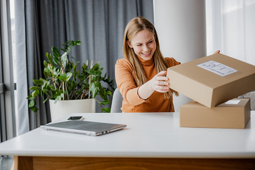 Smiling young woman preparing to open an online purchase package. She is sitting at the table. Cardboard boxes are on the table.