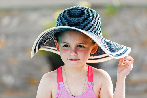 Portrait of a happy little blonde little girl in summer clothes and hat at public park she is sitting on grass