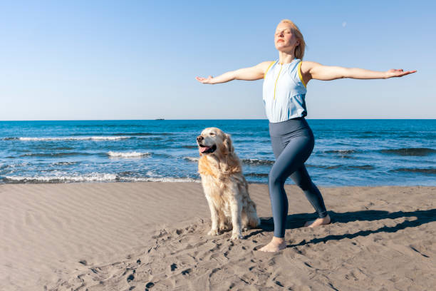 白人のアスレチックな女性は、太陽が彼女と彼女の美しい大きな犬の顔をしながら海辺のビーチでストレッチをすることで彼女の体をリラックスさせます。 - real people blue white friendship ストックフォトと画像
