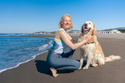 A beautiful white athletic girl with blond hair and her golden retriever dog enjoy a cuddle in the morning sun after training, on a sandy beach.