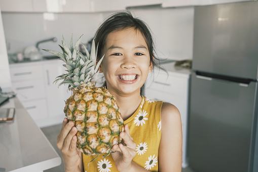 Happy young mixed Asian girl child holding fresh pineapple, healthy plant based vegan diet concept