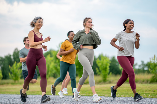 A large group of mature adults are seen from behind running together as part of a club.  They are dressed comfortably in athletic wear as they run down the road on a warm summer day.