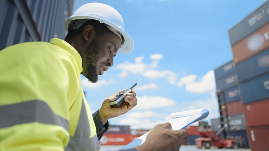 African American engineer or foreman wear hard hat safety vest, talking on the walkie-talkie, control cargo loading and shipping process at loading container yard for delivery to a cargo ship supporting the logistics.