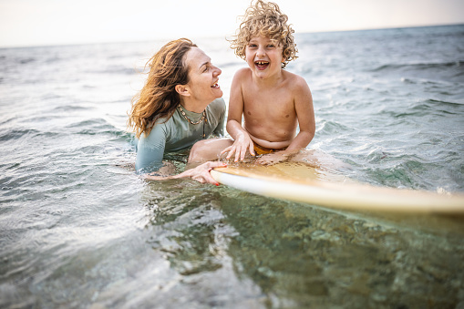 Photo of smiling boy having fun in the sea while floating on a surfboard with his mother.
