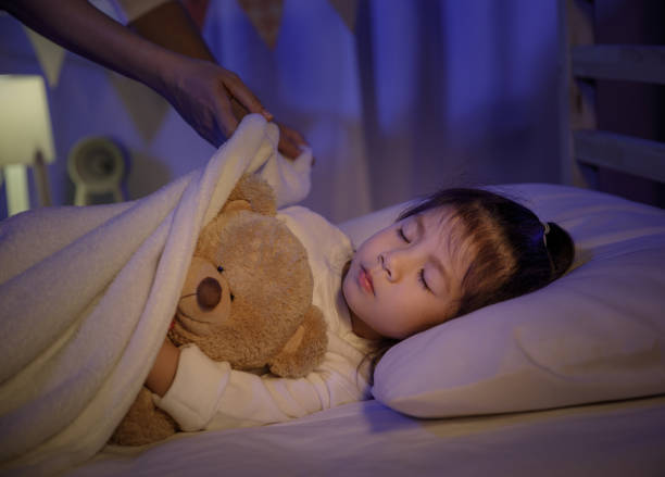 madre poniendo manta y acariciando la cabeza para su hija pequeña en la cama en un dormitorio oscuro por la noche, niña asiática abraza oso de peluche, niños cómodos en casa concepto - bedtime fotografías e imágenes de stock