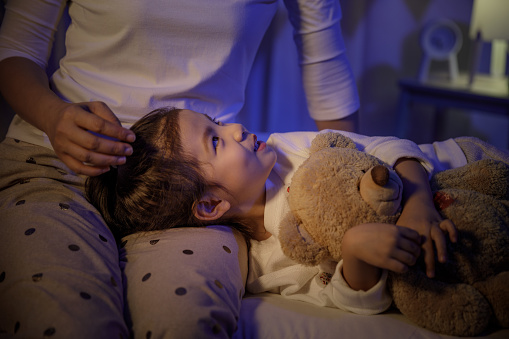 Mother stroking head of cute girl then sleeping on mom lap on bed in a dark bedroom at night, Child asian girl hug teddy bear, Comfortable children at home concept