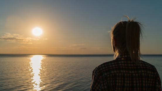 A young attractive woman is watching the sunset over the river. View from the back of a girl enjoying the beautiful scenery while traveling