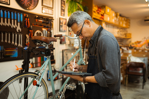 Senior man mechanic repairing bicycle brake at bicycle shop. He is wearing apron and using digital tablet.