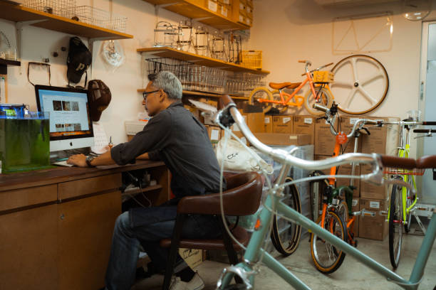 hombre asiático senior que trabaja en una computadora portátil en una tienda de bicicletas. - bicycle frame fotografías e imágenes de stock