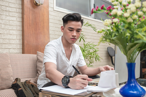 An asian freelance assistant or university student writes down some notes on a piece of paper. Working at home.