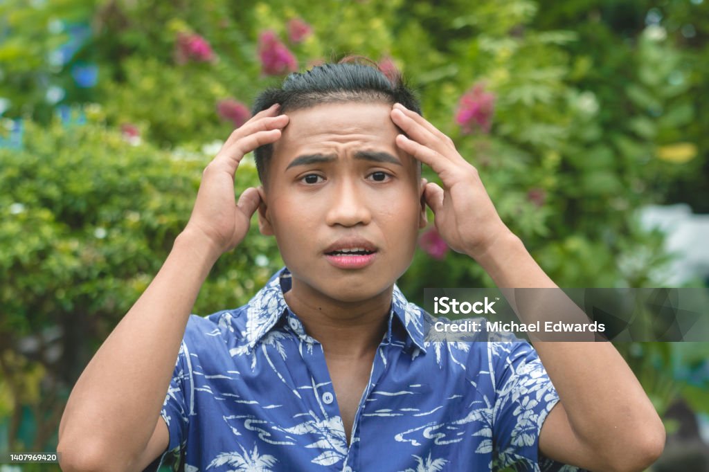 A panicked young Filipino man in a hawaiian shirt. Hands on forehead in shock. Outdoor garden scene. Shock Stock Photo