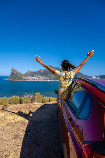 woman outside car window road trip with hands up, car at Chapmans Peak Drive in Cape Town South Africa looking out over ocean stock photo