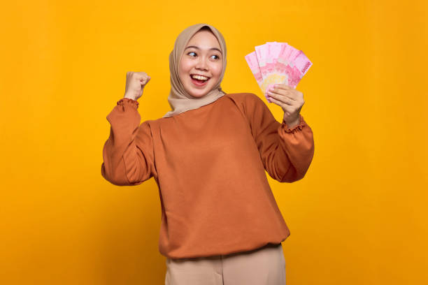 jeune femme asiatique excitée en chemise orange tenant des billets d’argent isolés sur fond jaune - indonesian currency photos et images de collection